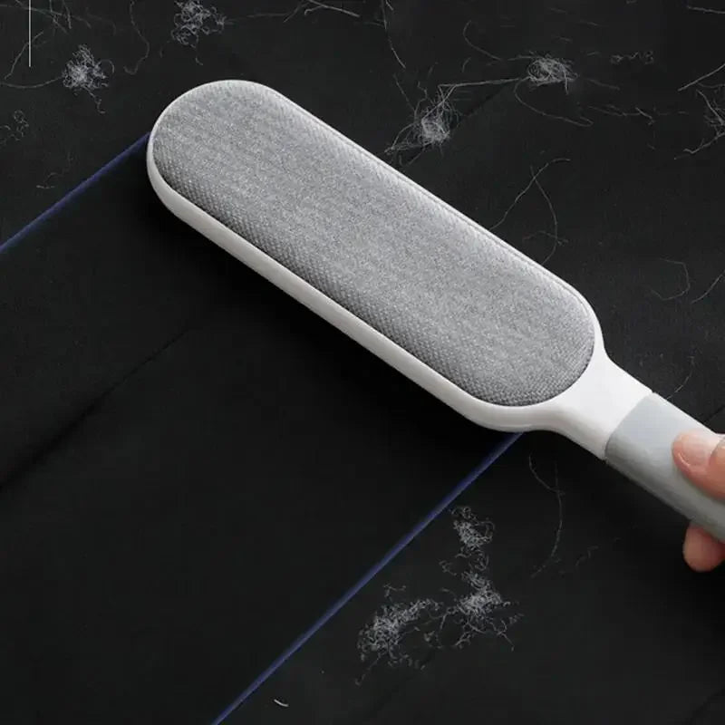 Double-sided grey plush pet hair removal brush cleaning fur from black fabric sofa and clothing.