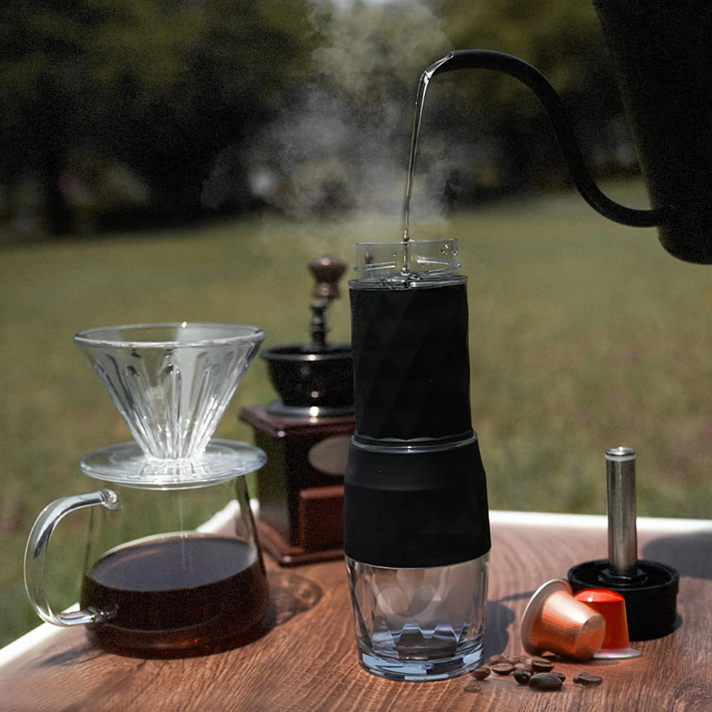 Portable black and glass coffee maker brewing with hot water on an outdoor table with a dripper and coffee beans.