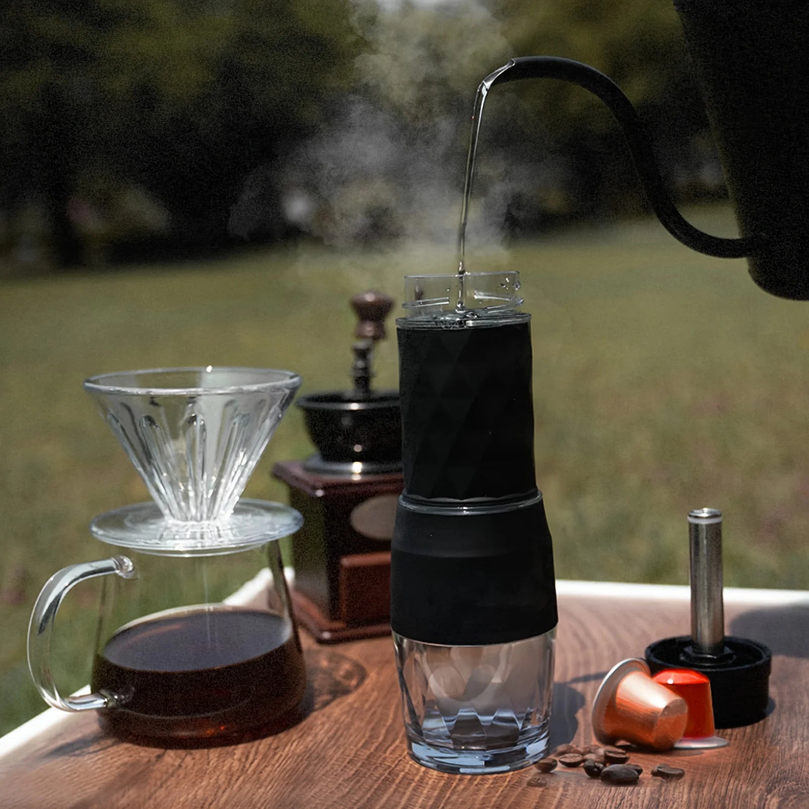 Portable black and glass coffee maker brewing with hot water on an outdoor table with a dripper and coffee beans.