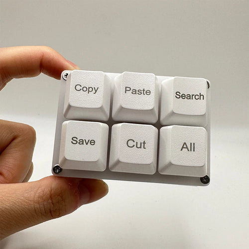 A hand holding a white, 6-key programmable mechanical macro keypad with shortcut labels on the keycaps.