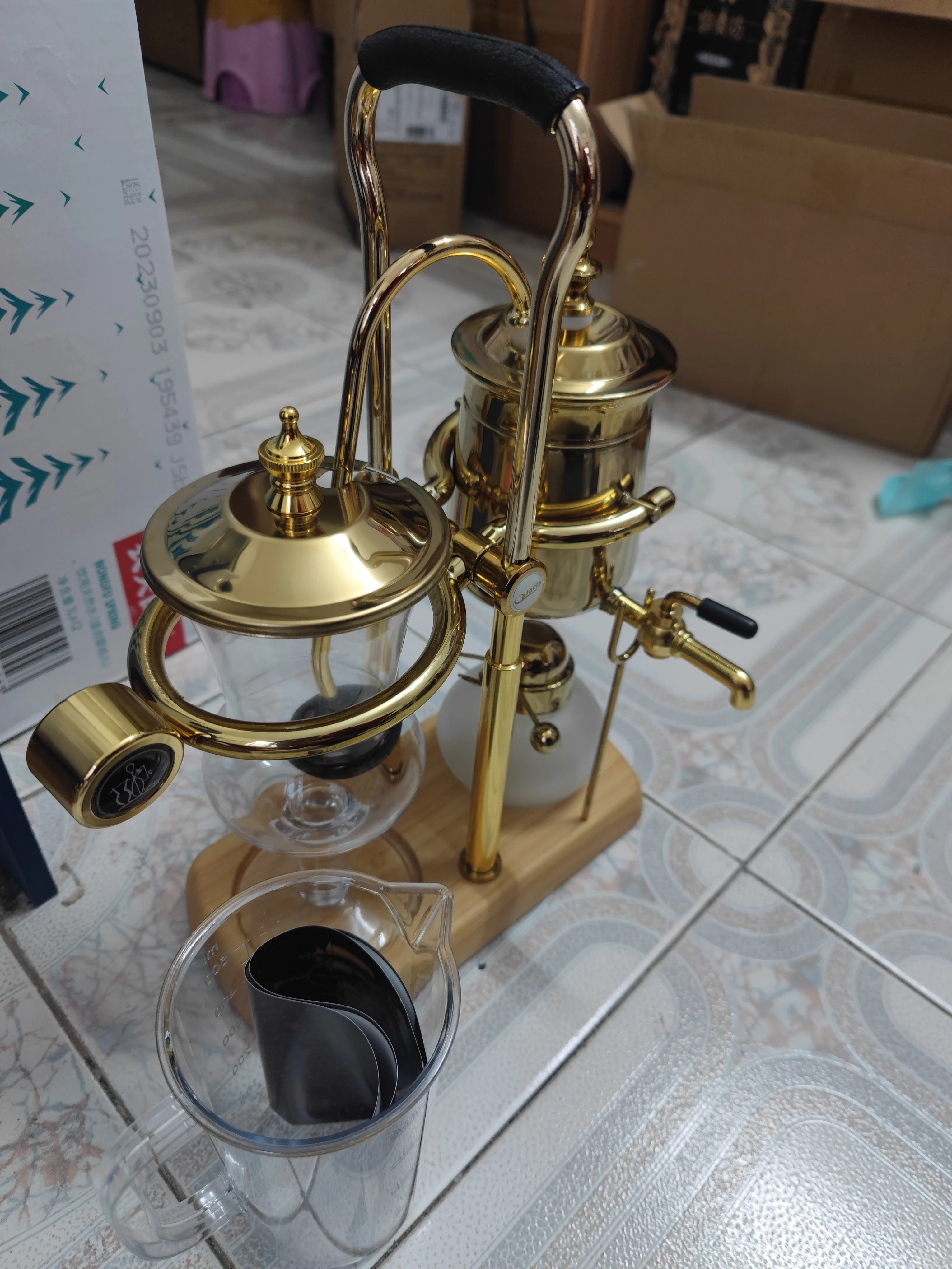 Gold balancing siphon coffee maker with glass chambers, metal finish, and wooden base stand for manual brewing.