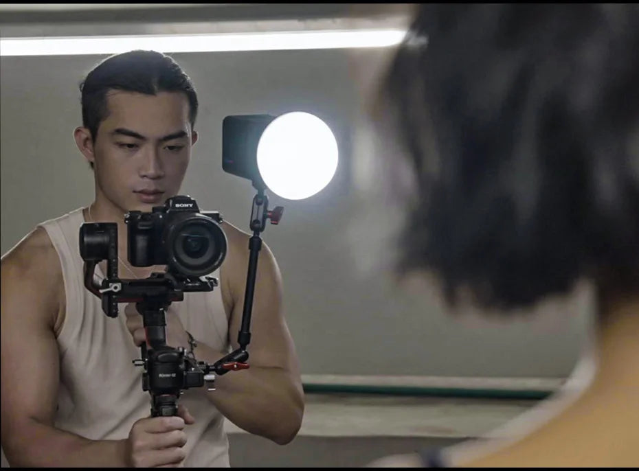 Black metal articulating arm mounting a round LED light to a camera gimbal stabilizer for video lighting.