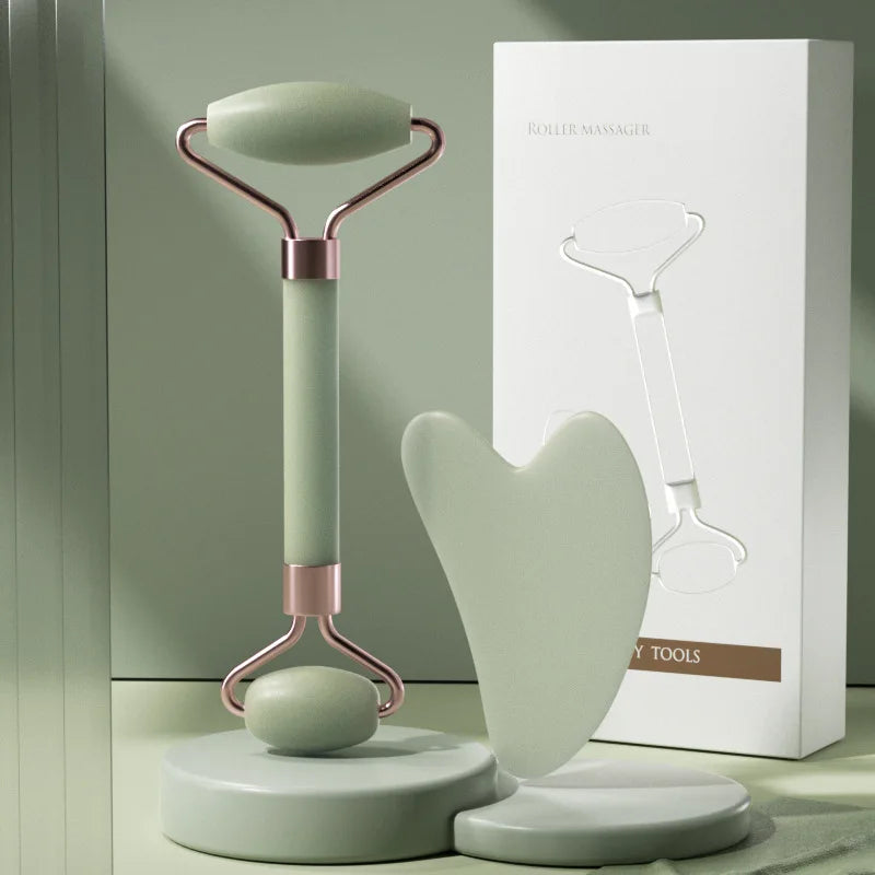 Sage green face roller and heart-shaped gua sha board on stands, with white product box in background.