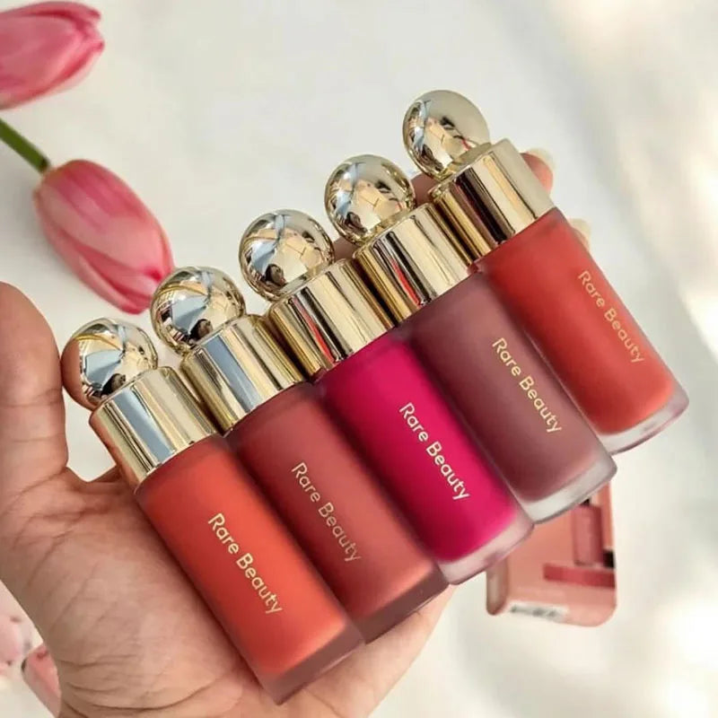Hand holding five shades of Rare Beauty Soft Pinch Liquid Blush in matte frosted glass bottles with gold caps.