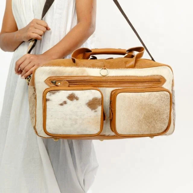 Western-style genuine hair-on-hide leather duffel travel bag with brown trim, exterior pockets, and shoulder strap.