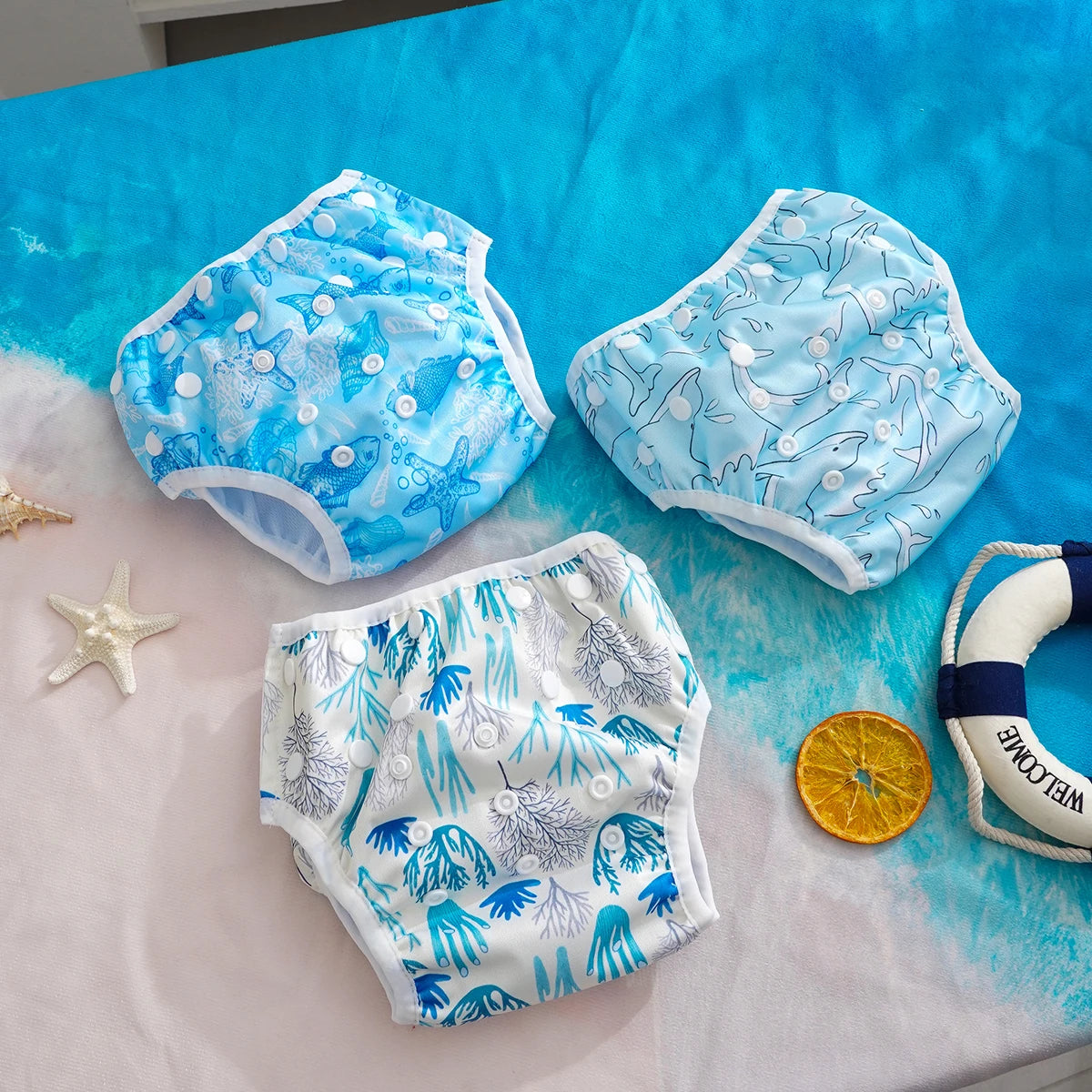 Set of three reusable baby swim diapers with ocean-themed starfish, shark, and coral patterns on a blue beach backdrop.