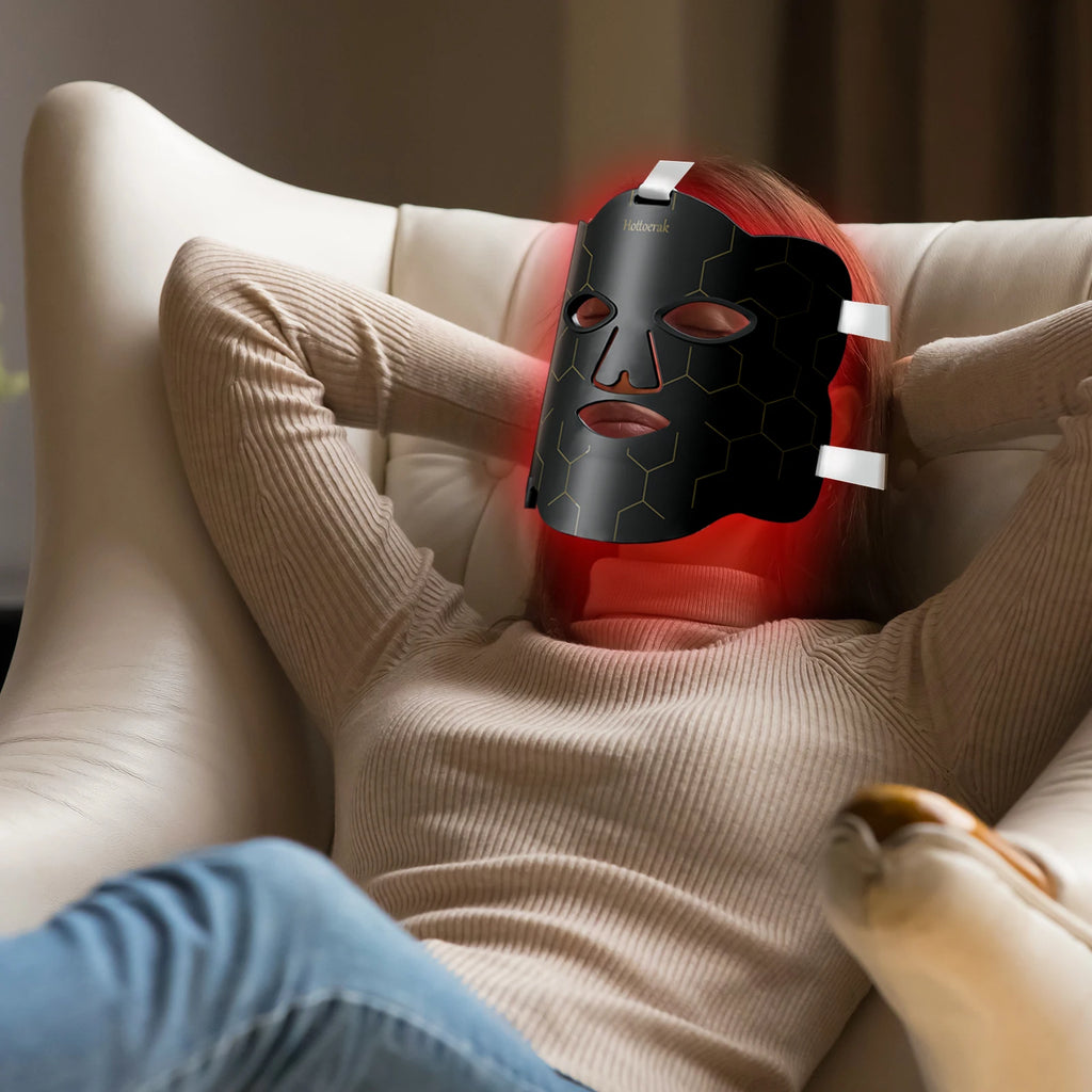 Woman relaxing in chair wearing a black Hottoerak flexible silicone LED mask for red light therapy skincare at home.