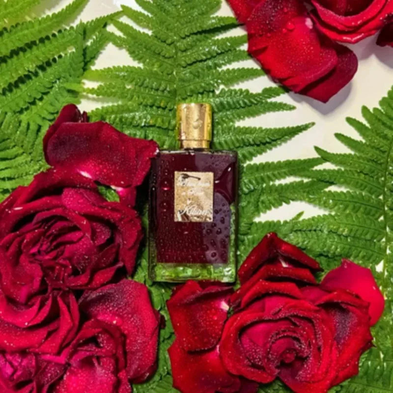 Rectangular glass perfume bottle with deep red liquid, gold cap, and label, surrounded by wet ferns and red roses.