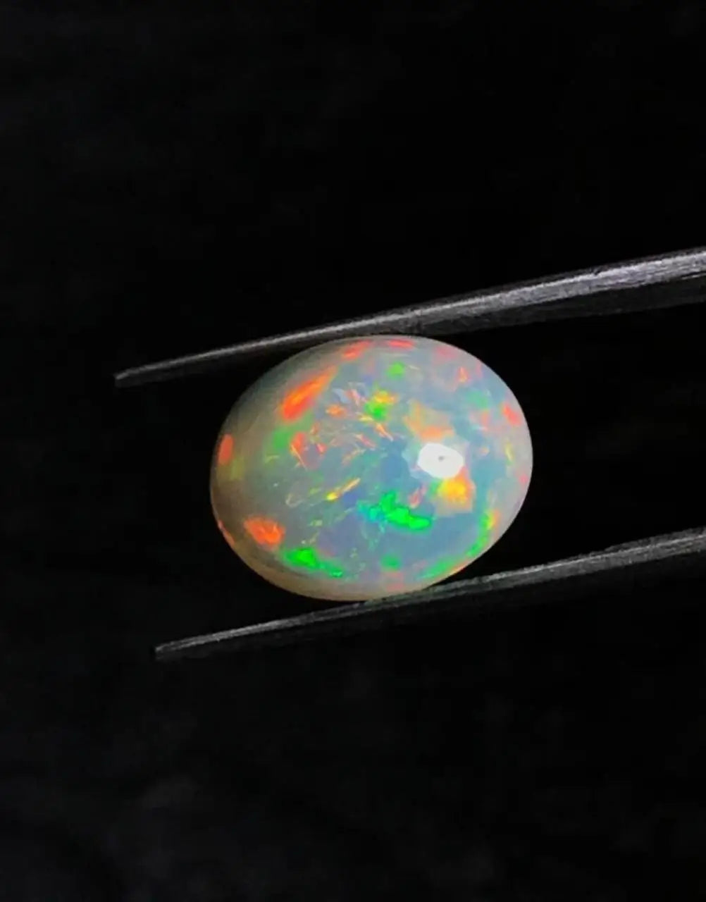 Oval opal cabochon with bright green fire and red-orange sparks between tweezers