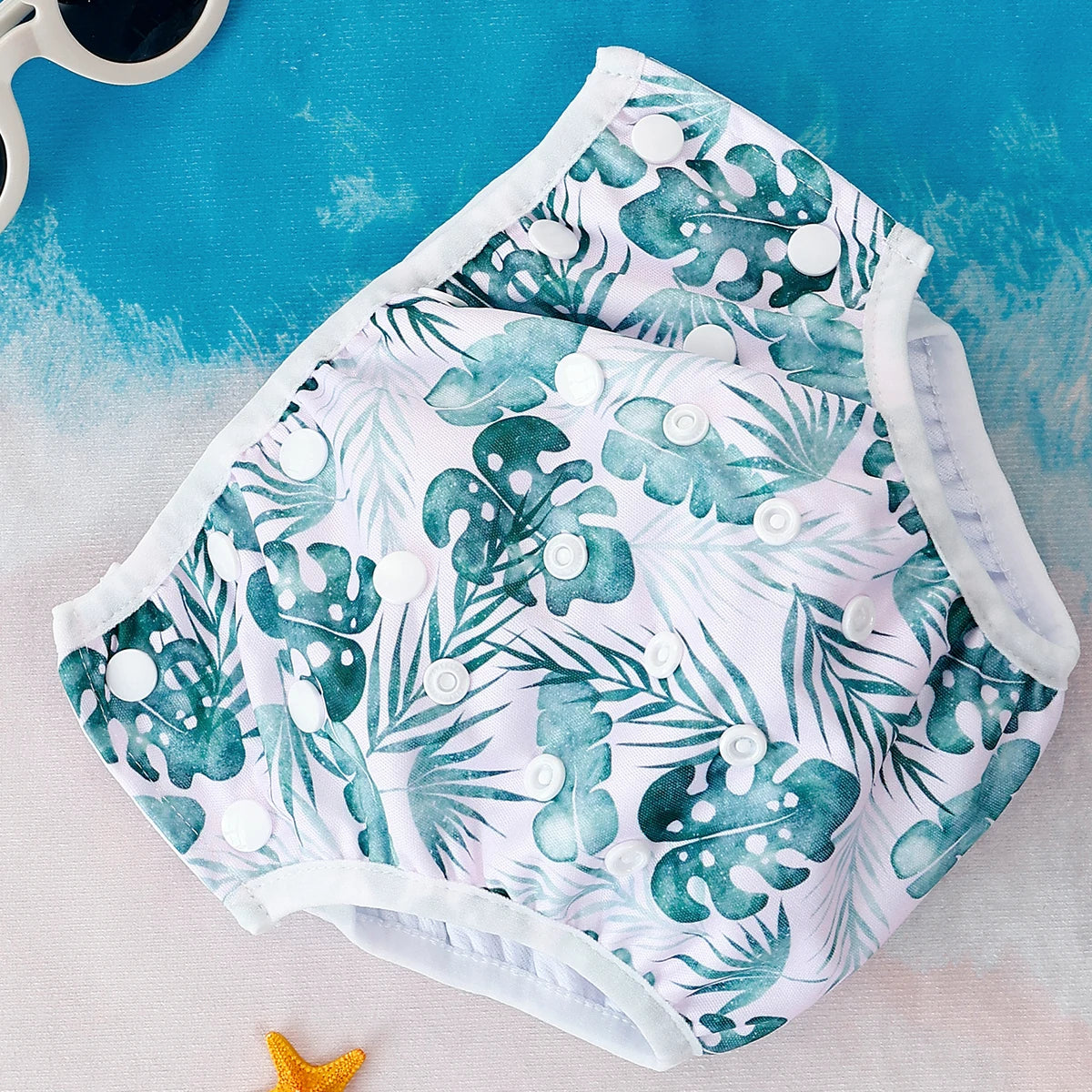 Reusable cloth diaper with tropical monstera leaf print and white snap buttons for adjustable baby care.
