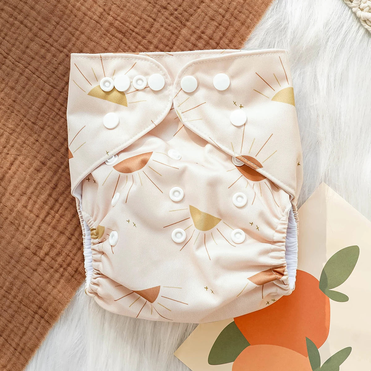 Reusable cloth baby diaper with tan sun print and white snap closures on a textured brown and white background.