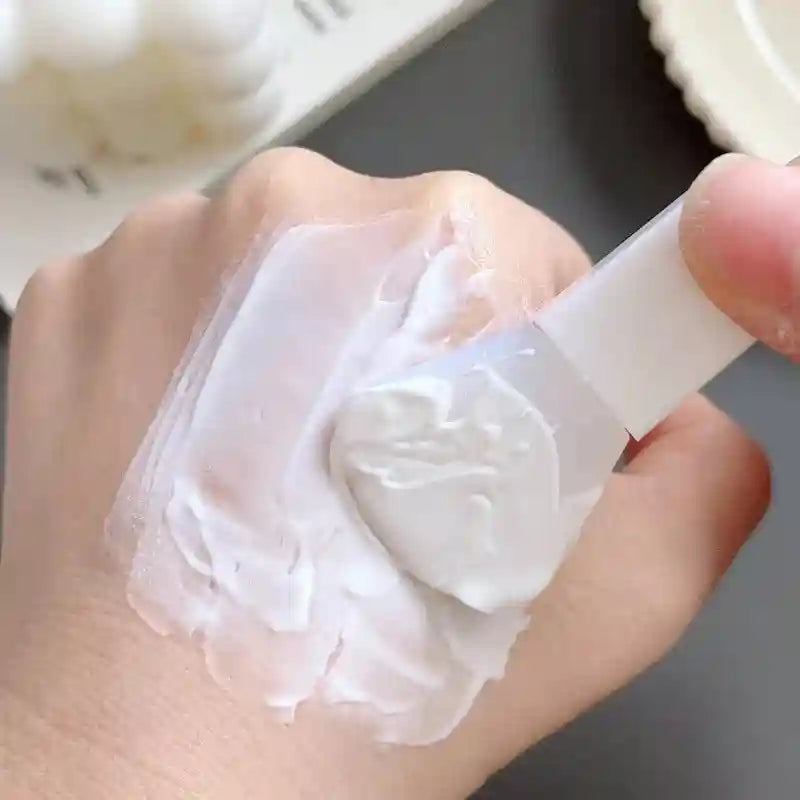 A hand using a flexible white silicone spatula to apply white cream onto the skin.