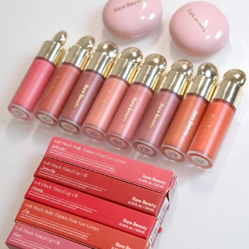 Rare Beauty Soft Pinch Tinted Lip Oil collection in various pink and peach shades with gold caps and red boxes.