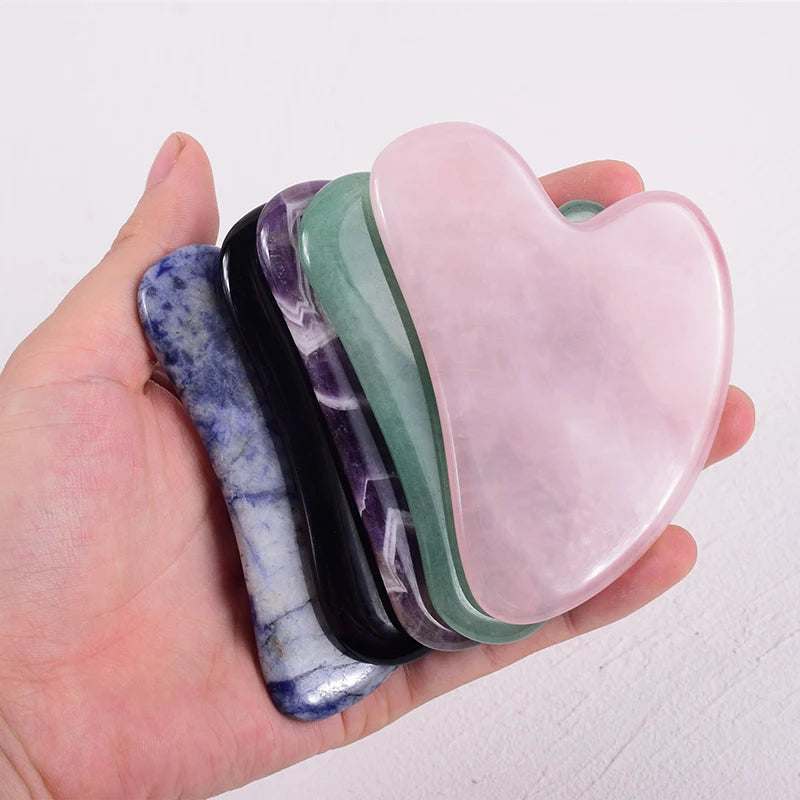 Hand holding a stack of heart-shaped Gua Sha facial massagers in various natural stone colors and textures.