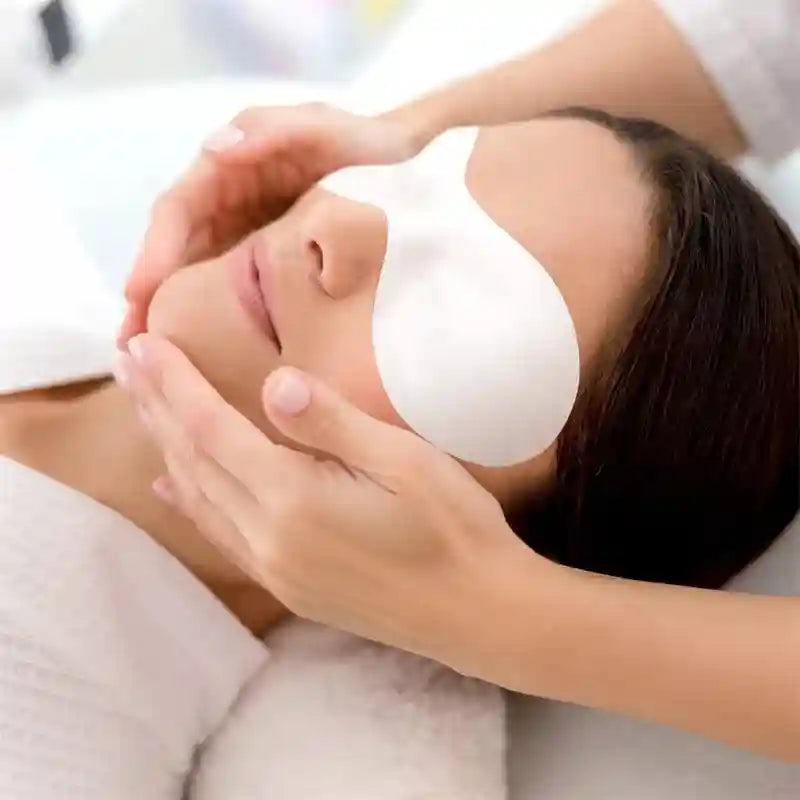 A person receives a facial with a white, disposable, crescent-shaped cotton eye mask covering their eyes.