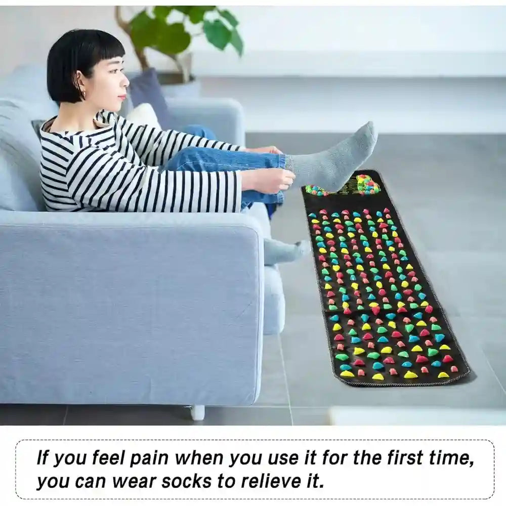 Woman using a black acupressure foot massage mat with colorful stone-like nodes for reflexology therapy on a floor.