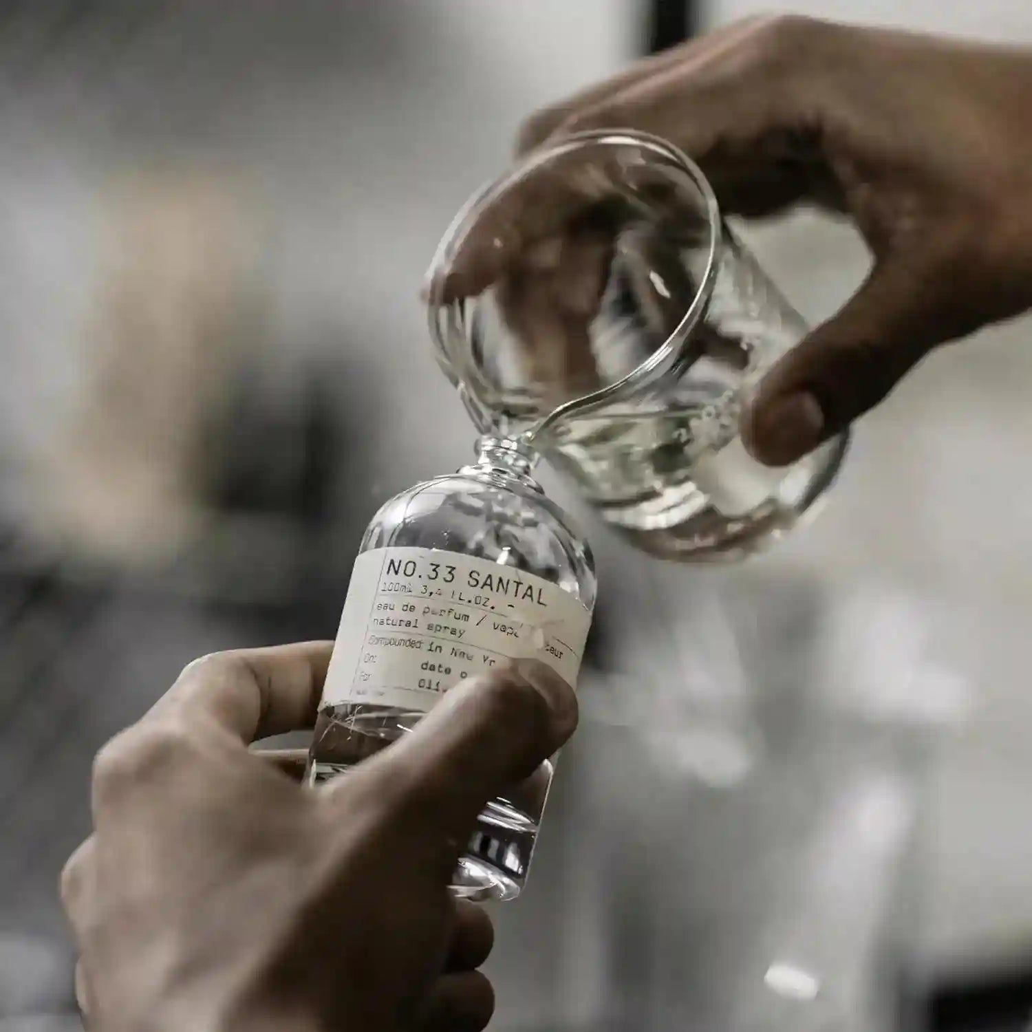 Hands pouring clear perfume from a glass beaker into a 100ml No.33 Santal glass fragrance bottle.