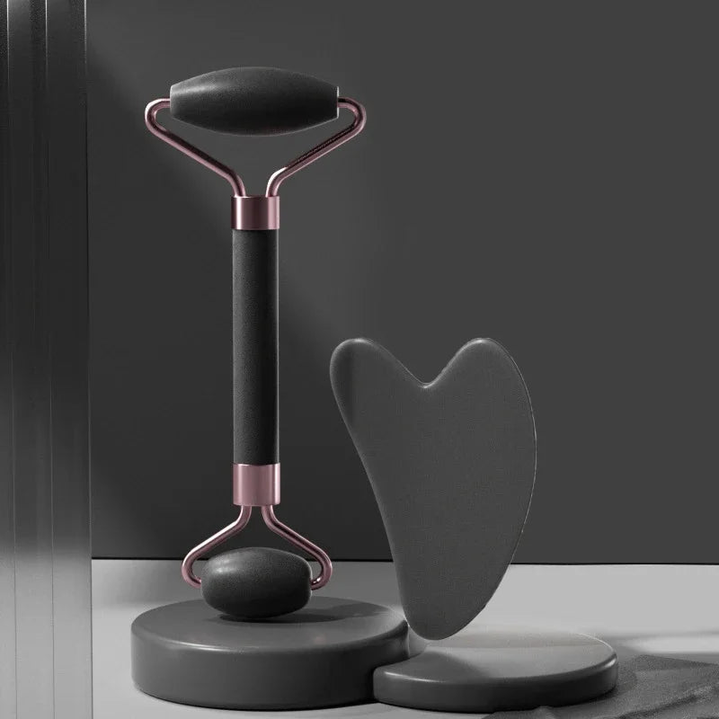 Matte black face roller with rose gold accents and a matching heart-shaped gua sha stone on grey display pedestals.