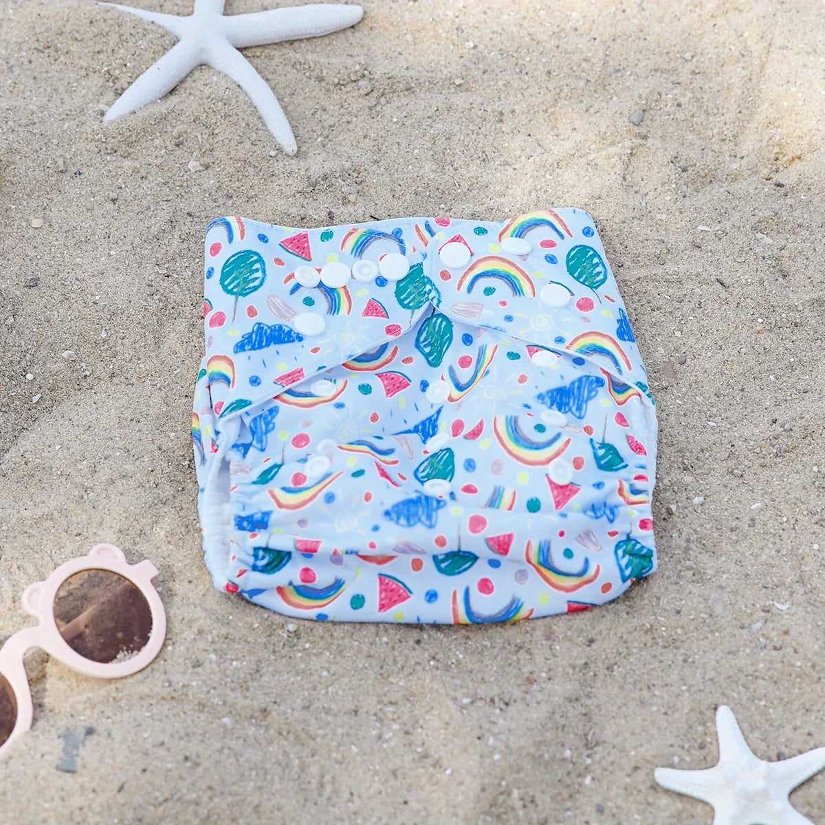 Reusable cloth baby diaper with rainbow and watermelon print, featuring snap closures on a sandy beach background.