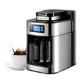Stainless steel digital coffee maker with glass carafe and programmable LCD display, showing a splash in a white cup.