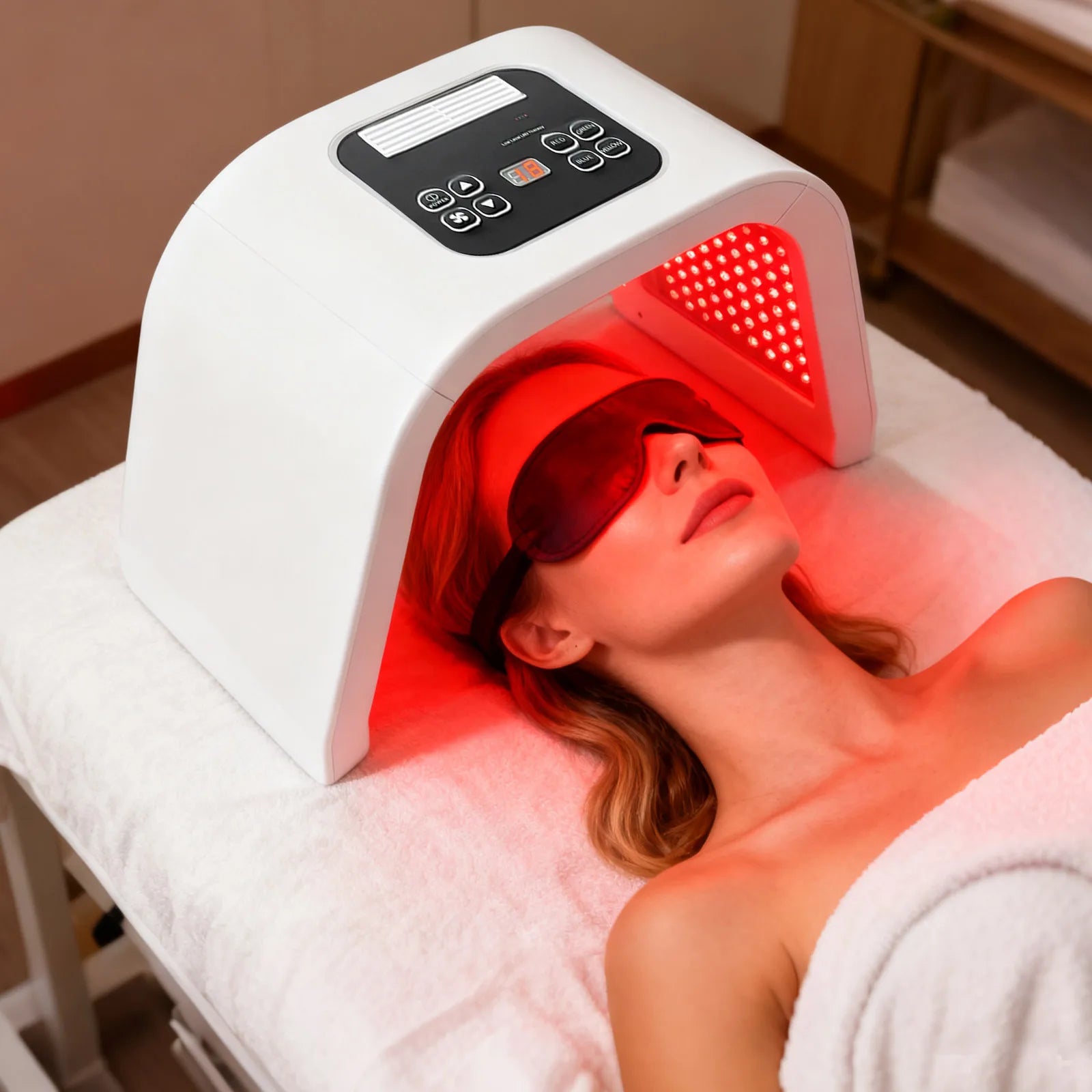 Woman in protective goggles lying under a white, arched LED light therapy machine emitting red light for facial care.