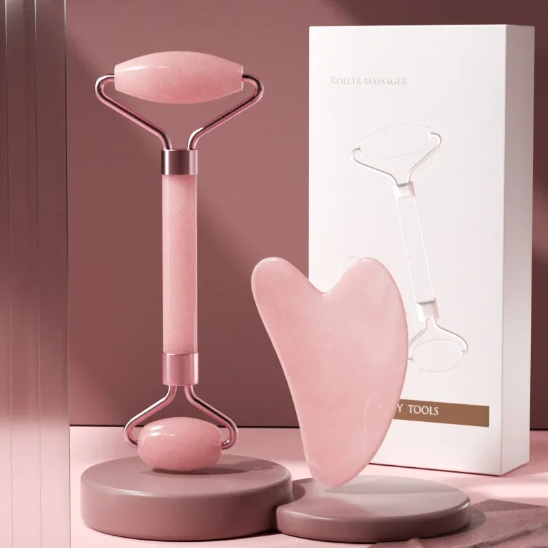 Pink resin face massage roller and heart-shaped gua sha board with gold accents, set next to white product packaging.
