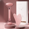 Pink resin face massage roller and heart-shaped gua sha board with gold accents, set next to white product packaging.