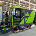 Electric street sweeper machine with a green and grey enclosed cab, front dual brushes, and warning light in warehouse.