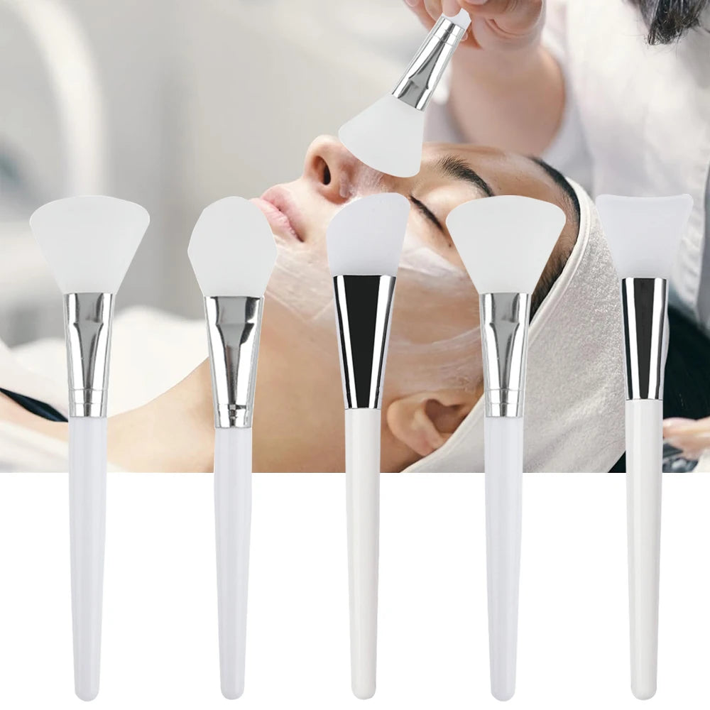 Assorted soft, white silicone face mask brushes with metallic ferrules and white handles against a spa background.