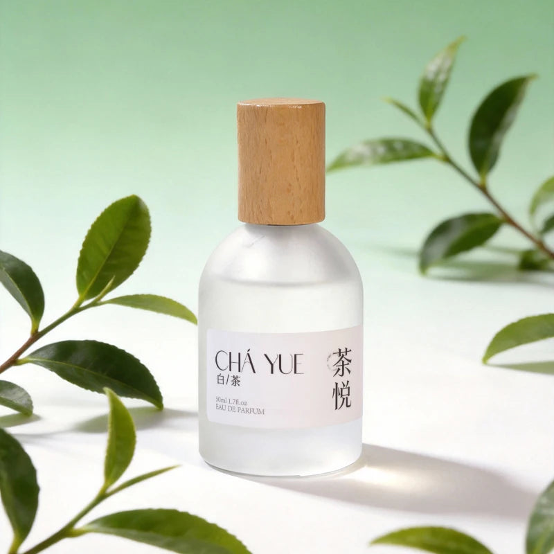 Frosted glass unisex white tea perfume bottle with wooden cap, surrounded by fresh green tea leaves on white background.