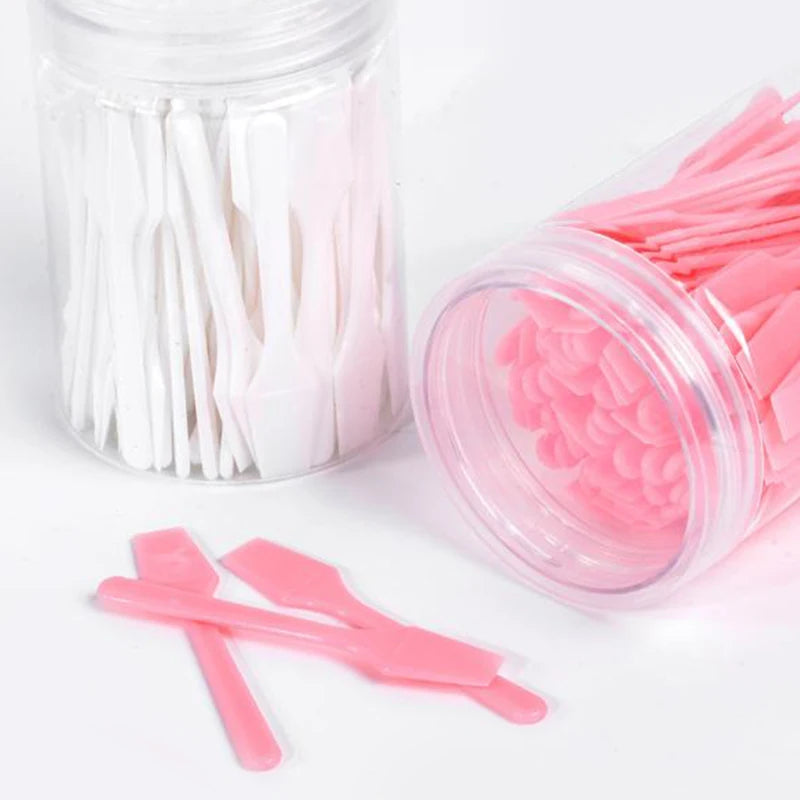 Two clear jars filled with small white and pink plastic cosmetic spatula scoops for applying creams and masks.