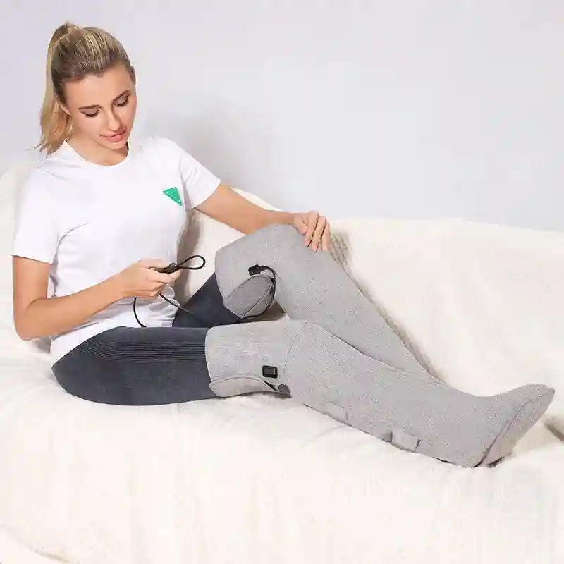 A woman sits on a white couch wearing tall, grey fabric leg-massage boots with an attached power controller.