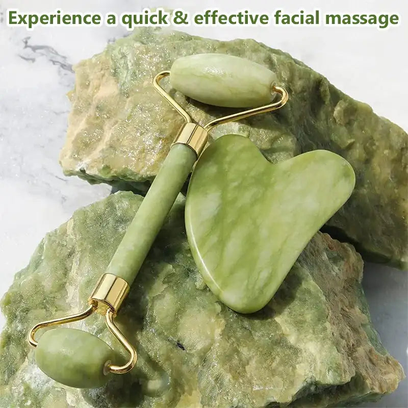 A green jade face roller and heart-shaped gua sha tool with gold metal accents, resting on a raw jade stone.