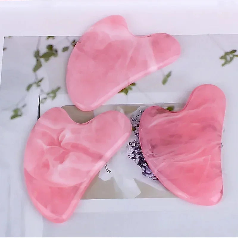 Three heart-shaped, pink marbled resin Gua Sha massage tools arranged on a white floral background.
