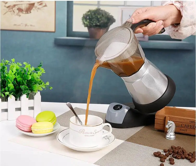Hand pouring coffee from a portable silver and black electric moka pot maker into a white ceramic teacup.