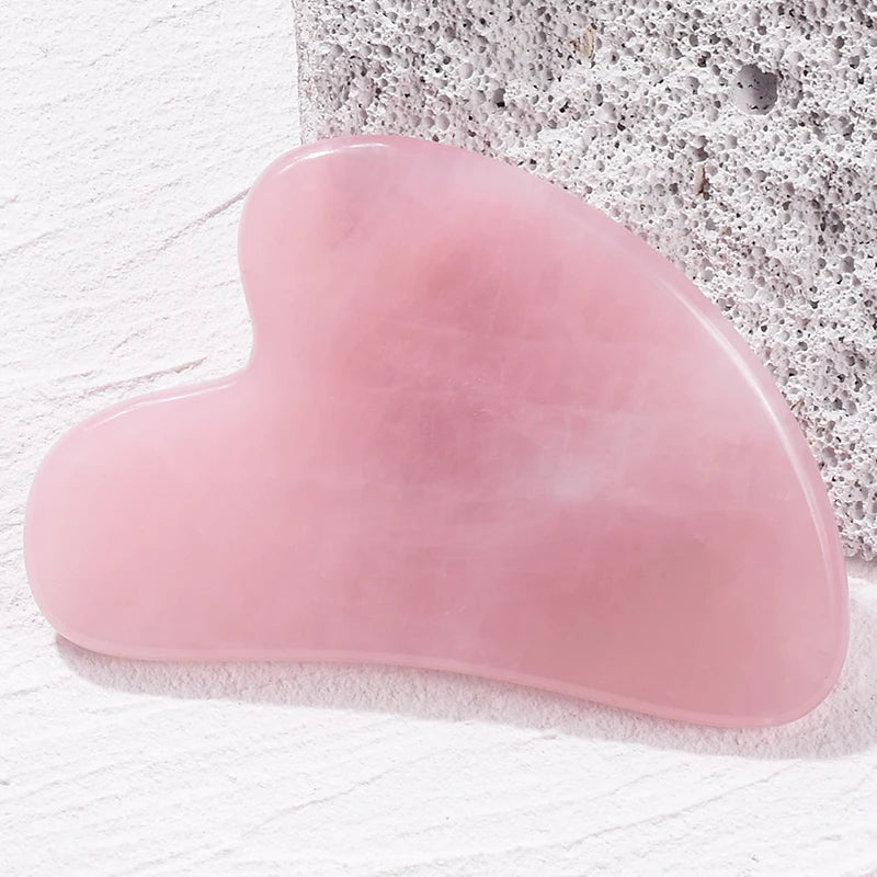 Heart-shaped light pink rose quartz Gua Sha massage tool resting on a textured white surface.