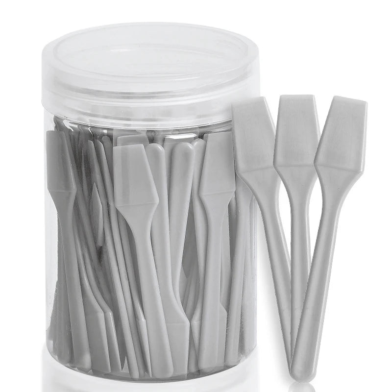 A clear plastic jar filled with 100 small, grey plastic cosmetic spatulas for applying creams and masks.