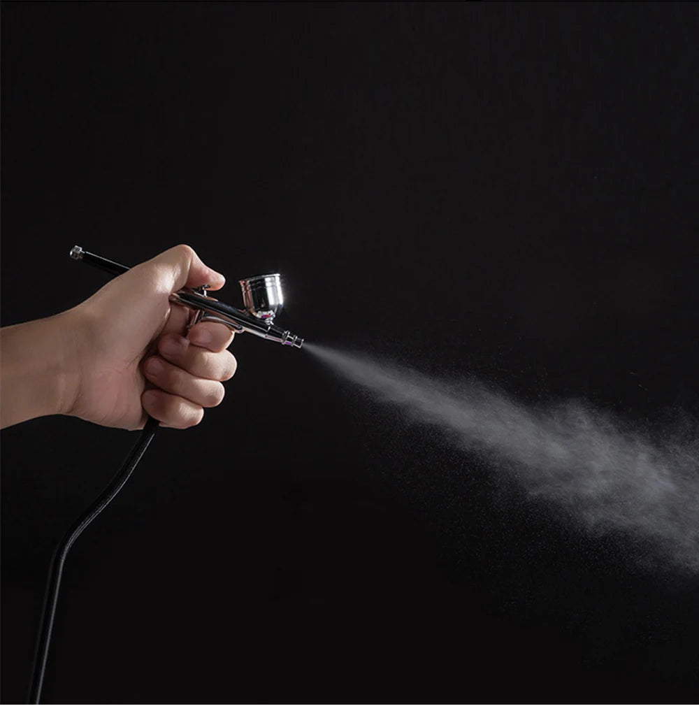 A hand holding a metallic silver airbrush nozzle, spraying a fine mist against a plain black background.