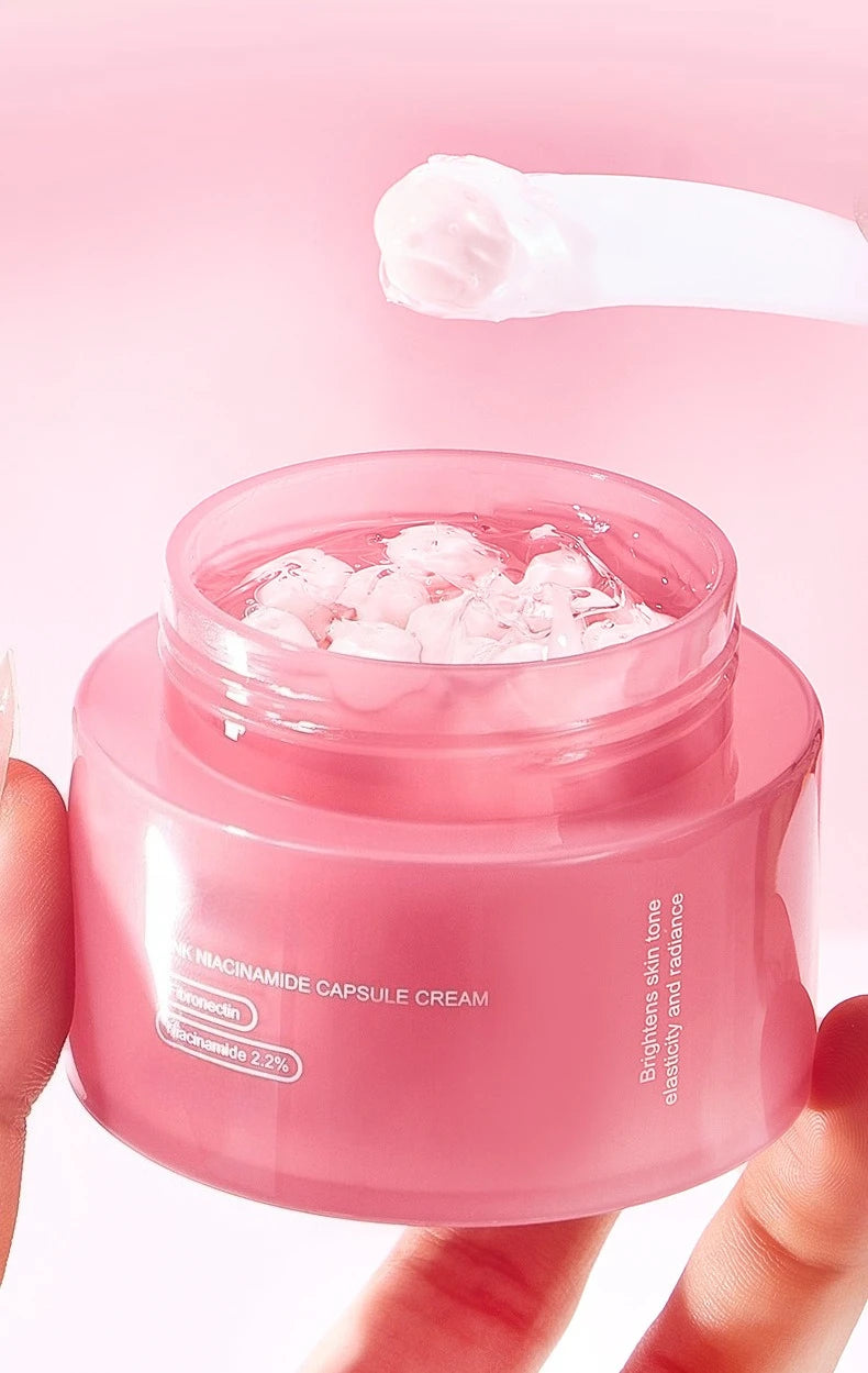 A round pink jar of face cream containing visible pink capsules, with a scoop of cream being lifted above it.