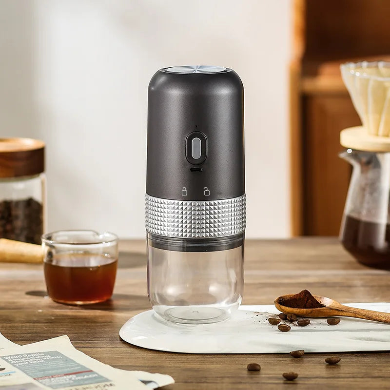 Portable electric coffee grinder, matte black and silver design with a clear container on a wooden kitchen table.