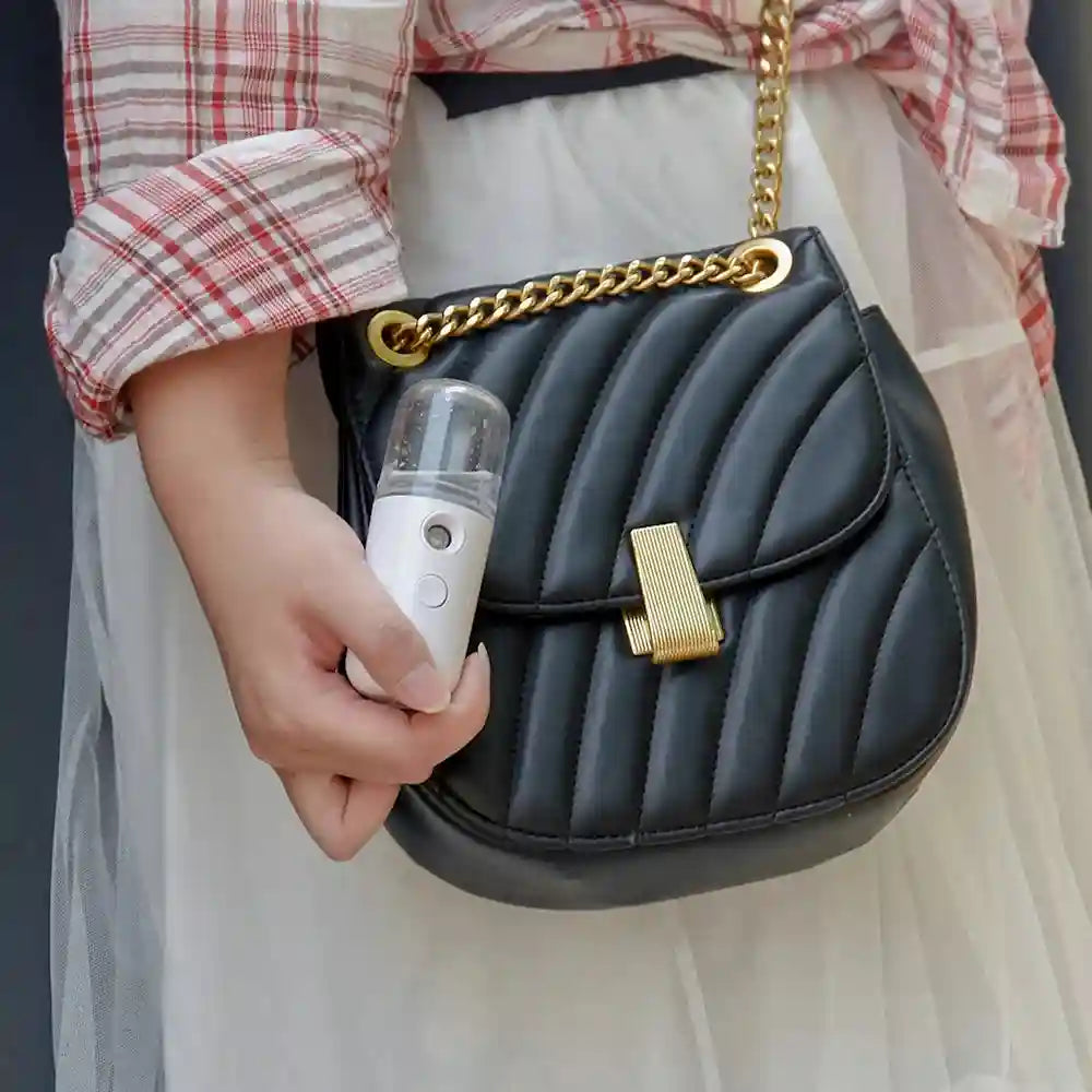Portable white nano facial mister sprayer for hydrating skin, held in hand next to a black quilted designer handbag.