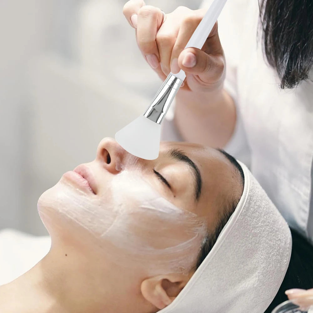 A person uses a white, flexible silicone spatula brush to apply a creamy facial mask to a woman's skin.