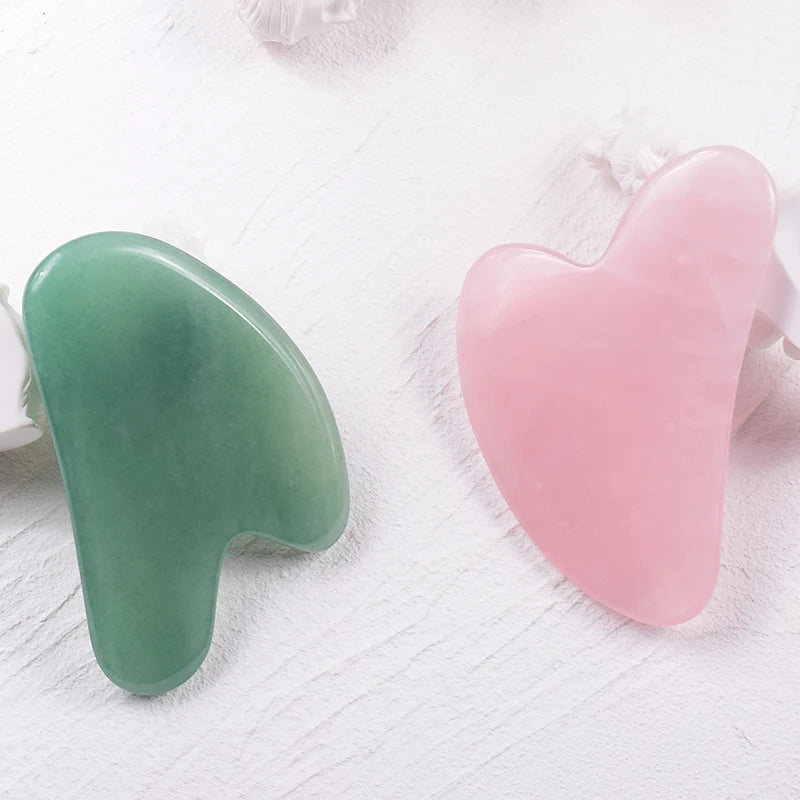 Two heart-shaped gua sha facial massage tools, one in green jade and one in pink rose quartz, on a white background.