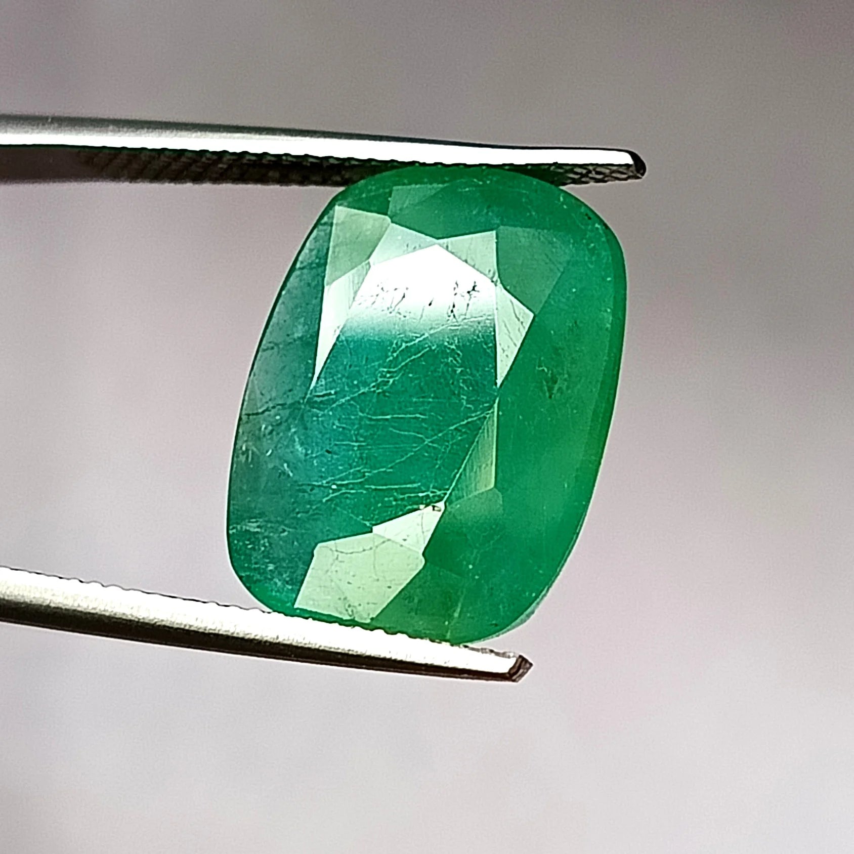 Green oval emerald gemstone, faceted front view, angled in metal tweezers.