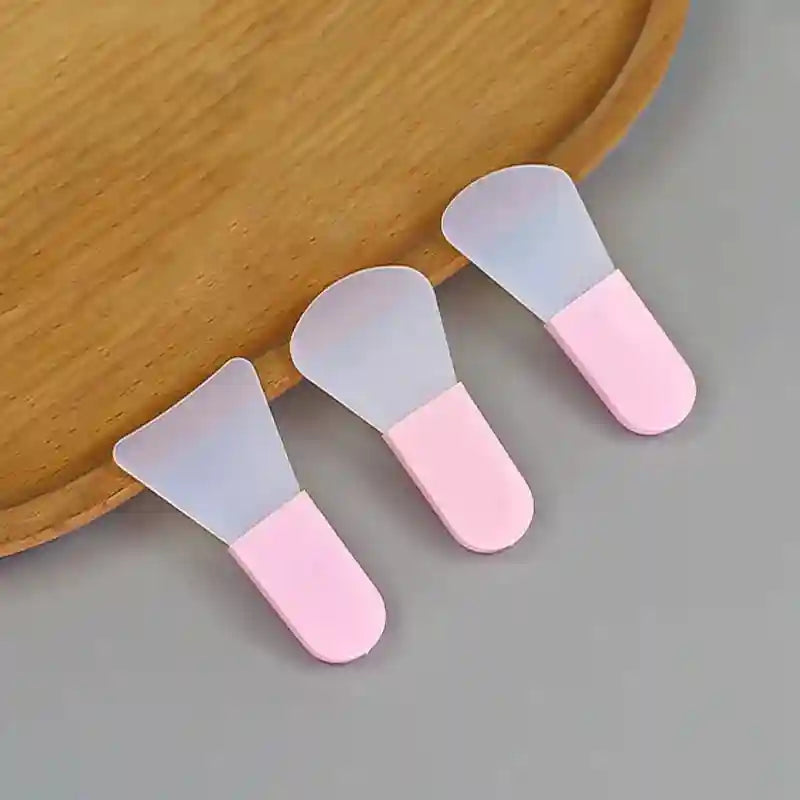 Three silicone facial mask brushes with translucent flexible heads and opaque pastel pink handles on a wooden tray.