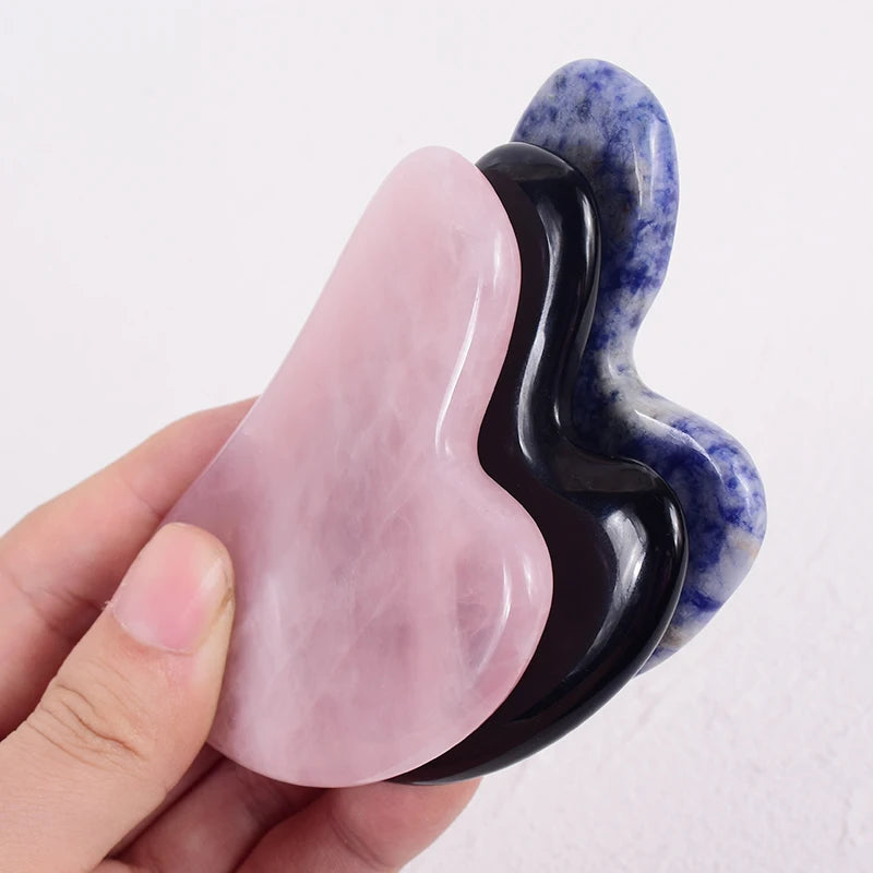Hand holding three heart-shaped Gua Sha facial massage tools made of pink rose quartz, black obsidian, and blue stone.
