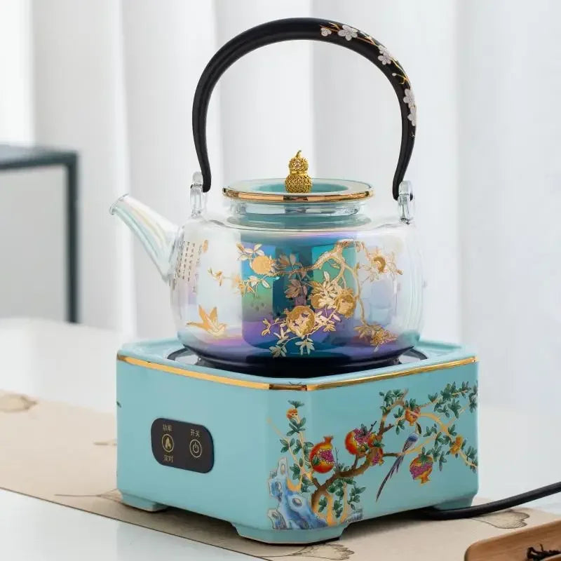 Elegant glass tea kettle with gold floral print on a decorative blue ceramic electric warming base.