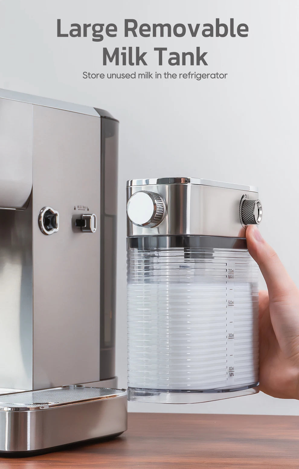 Hand holding a removable 700ml clear milk tank for a stainless steel espresso coffee machine on a wooden table.