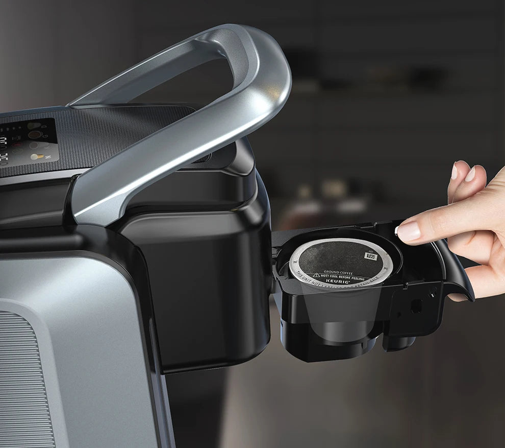 Hand inserting a K-Cup pod into a sleek black and silver Keurig single-serve coffee maker with a metallic handle.