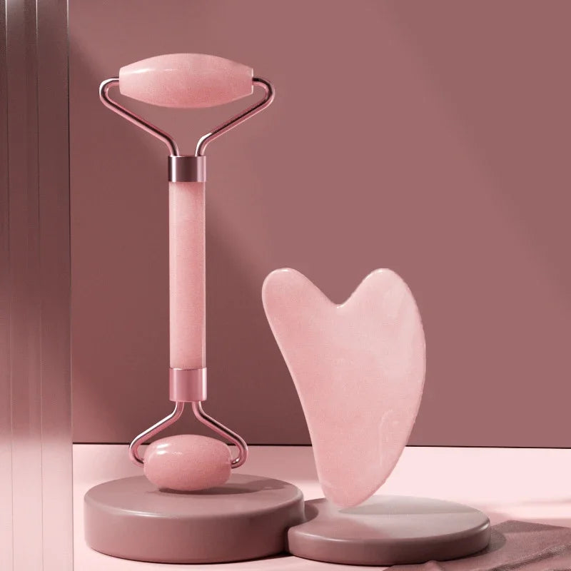 Pink resin facial roller and heart-shaped gua sha board on minimalist pink pedestals.